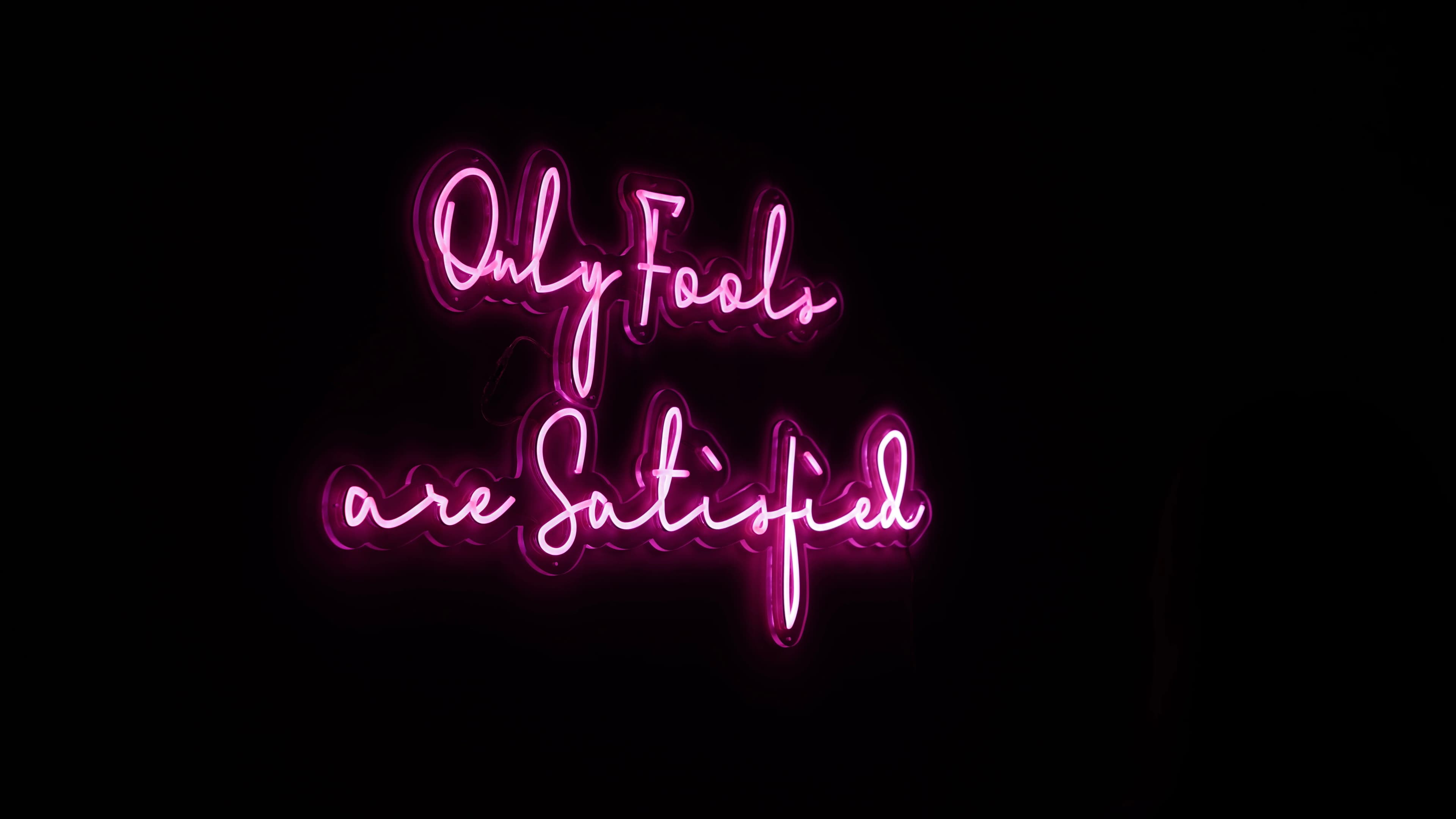 A pink neon sign reading "Only Fools are Satisfied" glowing on a dark wall